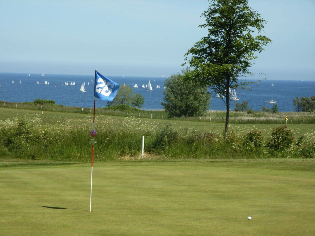 Ein Golfball kurz vorm einlochen. Im Hintergrund sieht man die Ostsee mit ihren Segelbooten